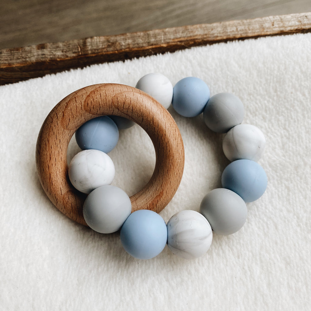 Baby teether in light blue, white and grey with a wooden ring.