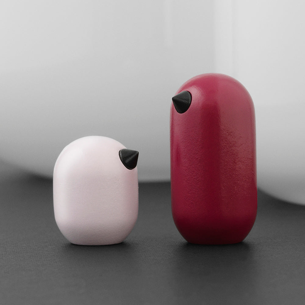 Two small wooden birds, one pink and one magenta, on a dark surface with a light grey background.