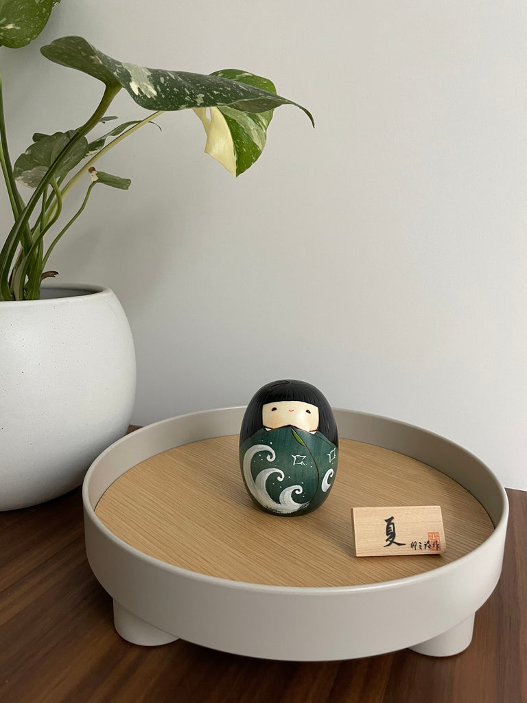 wooden kokeshi doll with wave detail on body in dark green on decorative tray next to plant
