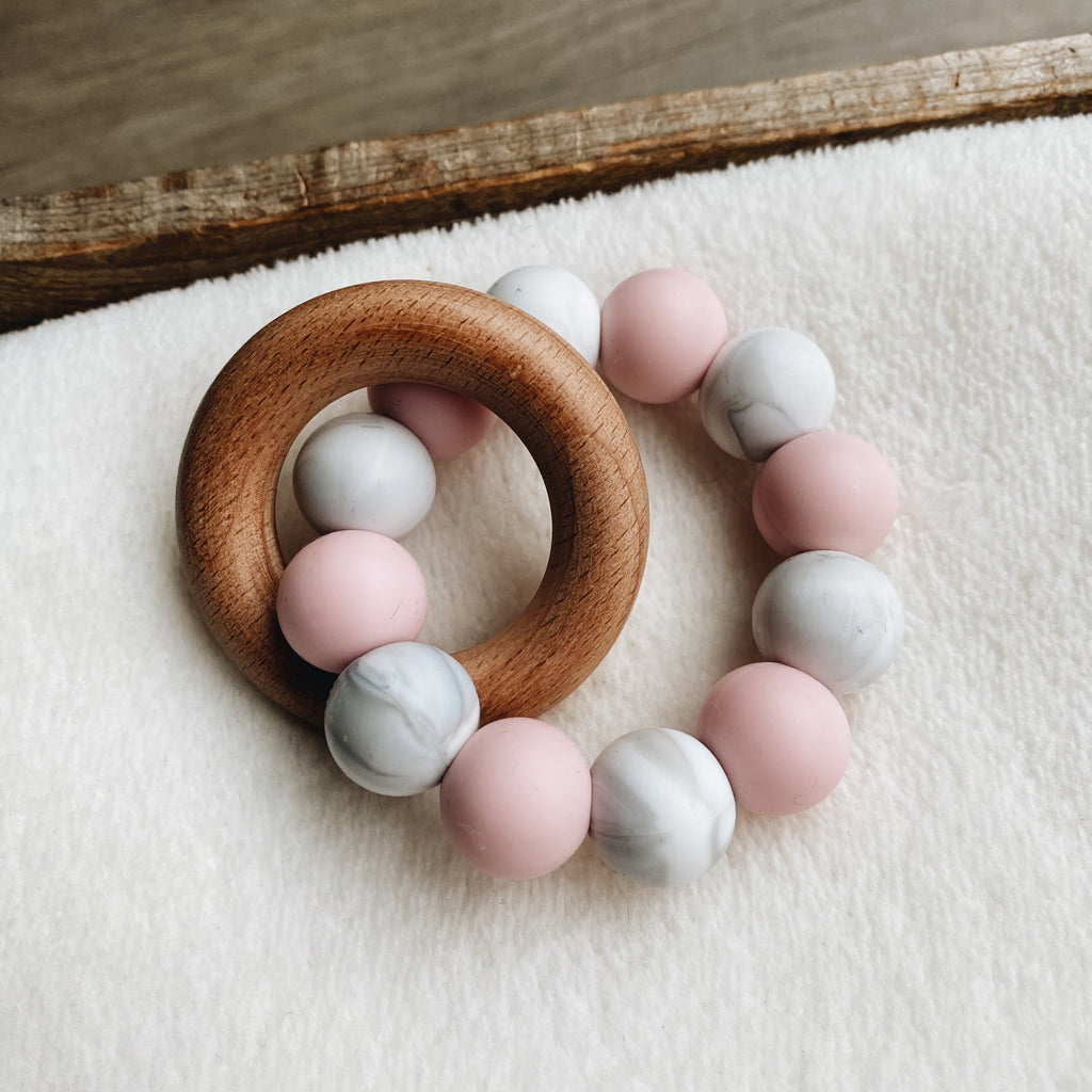 Baby teether in baby pink, white and grey with a wooden ring.